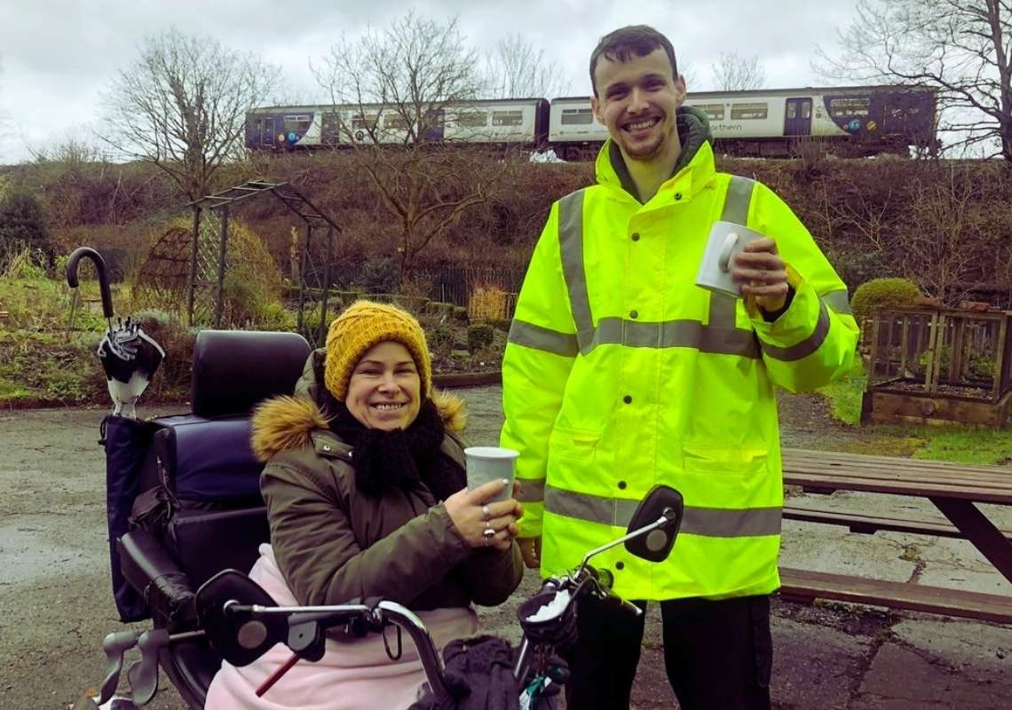 A man and woman smile holding cups of tea. They are in a community garden. The woman is in a wheelchair and wearing a wooly hat. The man is wearing a high viz jacket.