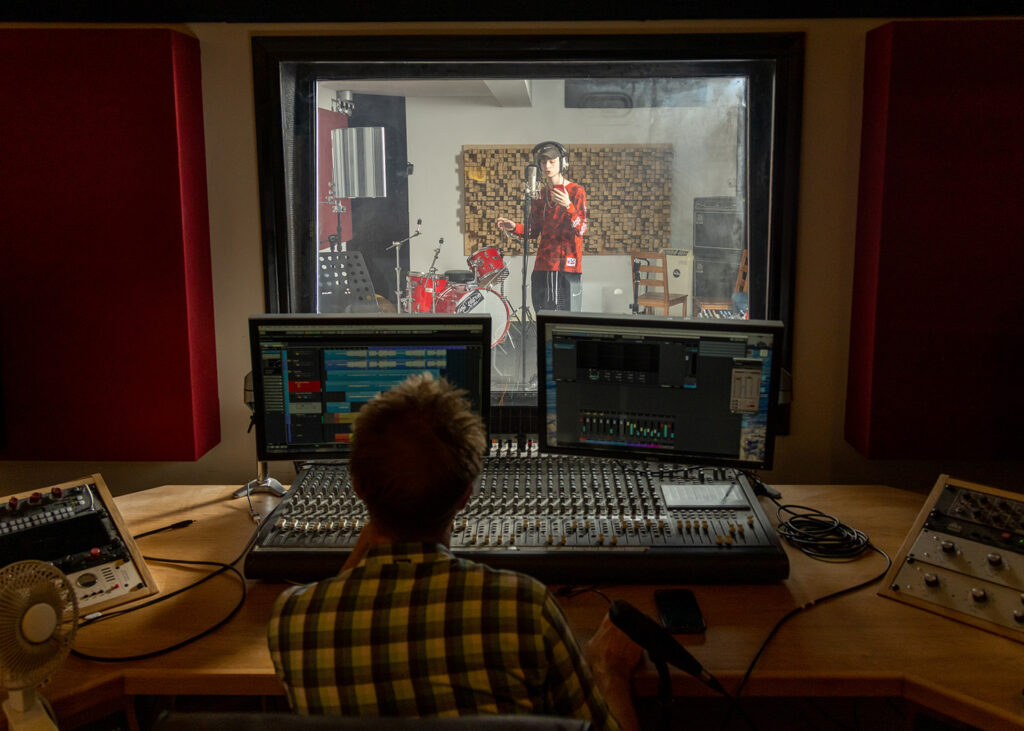 Jake in the studio rapping into the mic in the background. With the music coach sitting at the foreground in front of various music production equipment.