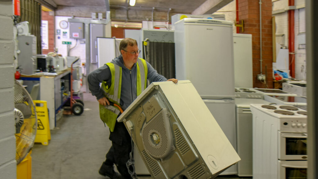 Man in high-vis vest wheels a washing machine on a trolley through a workshop full of other white-goods.