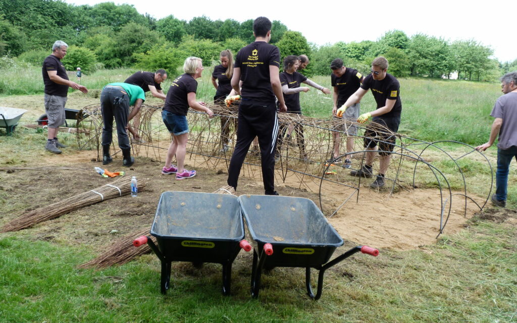 Group of people in black branded T-shirts working outdoors, with wheelbarrows in foreground