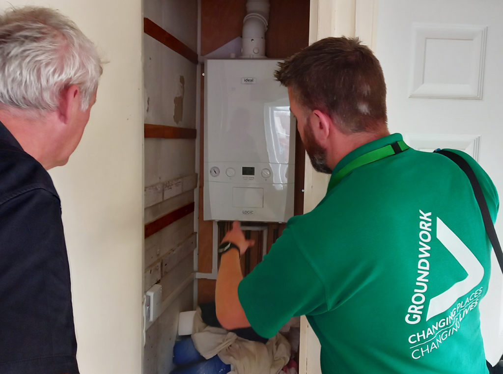 Man wearing green Groundwork shirt pointing at boiler with another man watching.
