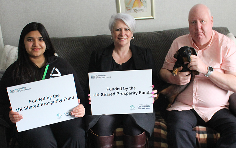 Two women and a man sitting on sofa, the women are holding white papers with the text "Funded by the UK Shared Prosperity Fund". The man holds a small dog.