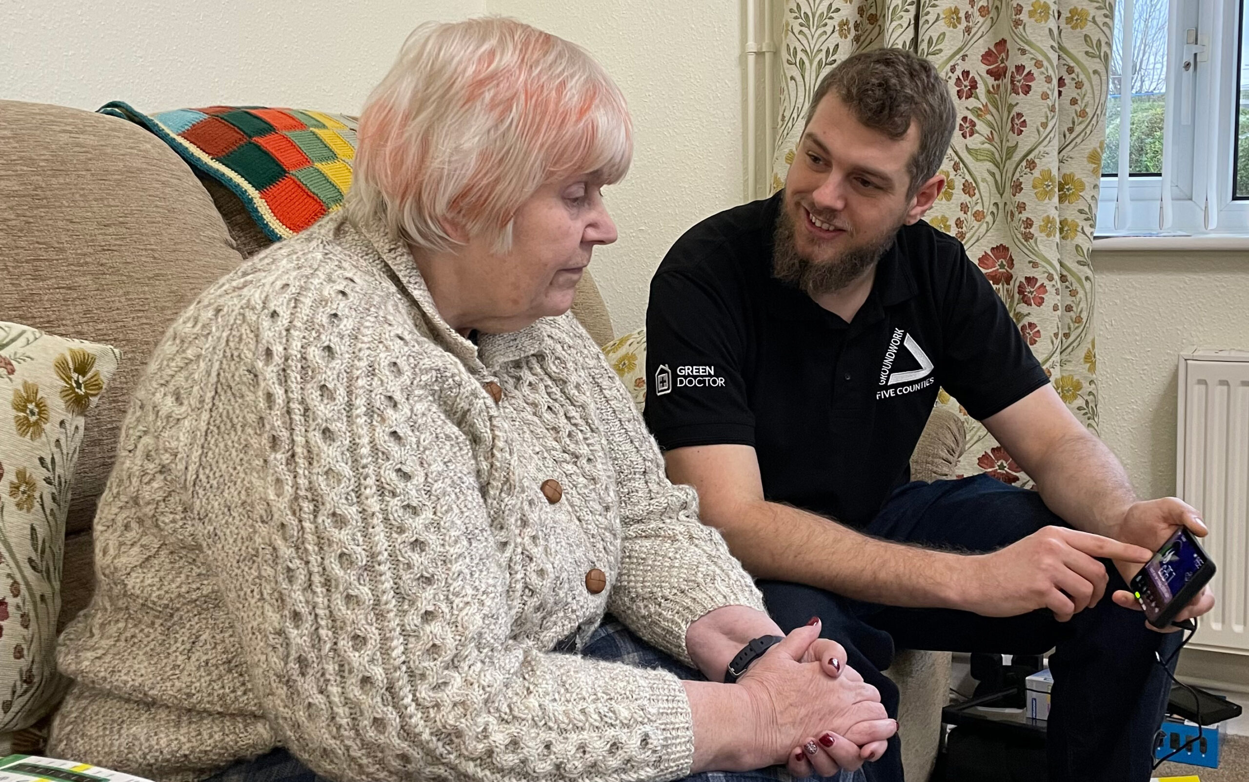 Elderly woman wearing grey cardigan and young man in black T-shirt with Green Doctor logos looking at smart meter.