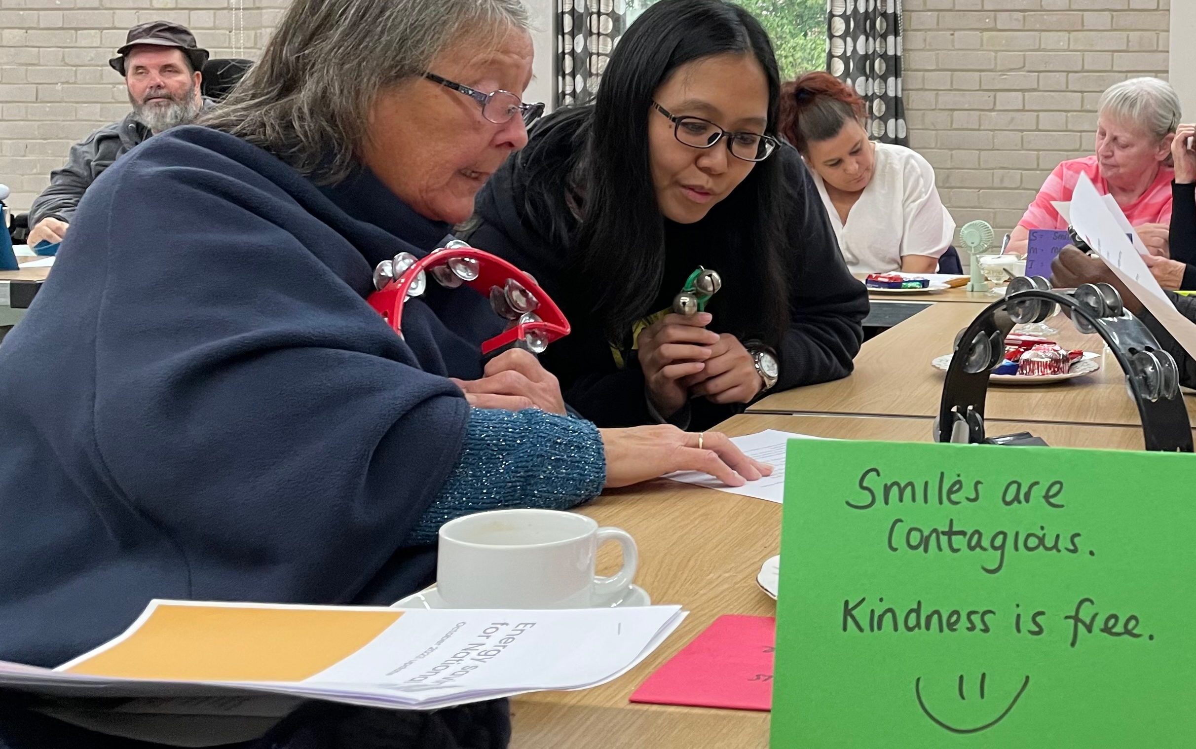 Elderly Caucasian woman and young Asian woman holding musical instruments sitting at table. Green card with black text "smiles are contagious. kindness is free" with smiley face.