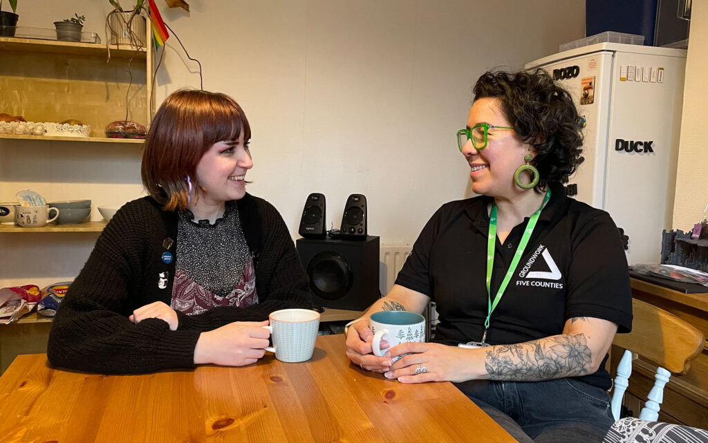 Two women sitting at kitchen table with cups, smiling at each other.