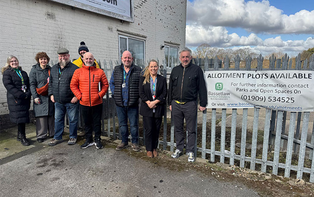 Group of people standing by metal gates with white banner with black text "Allotment Plots available".