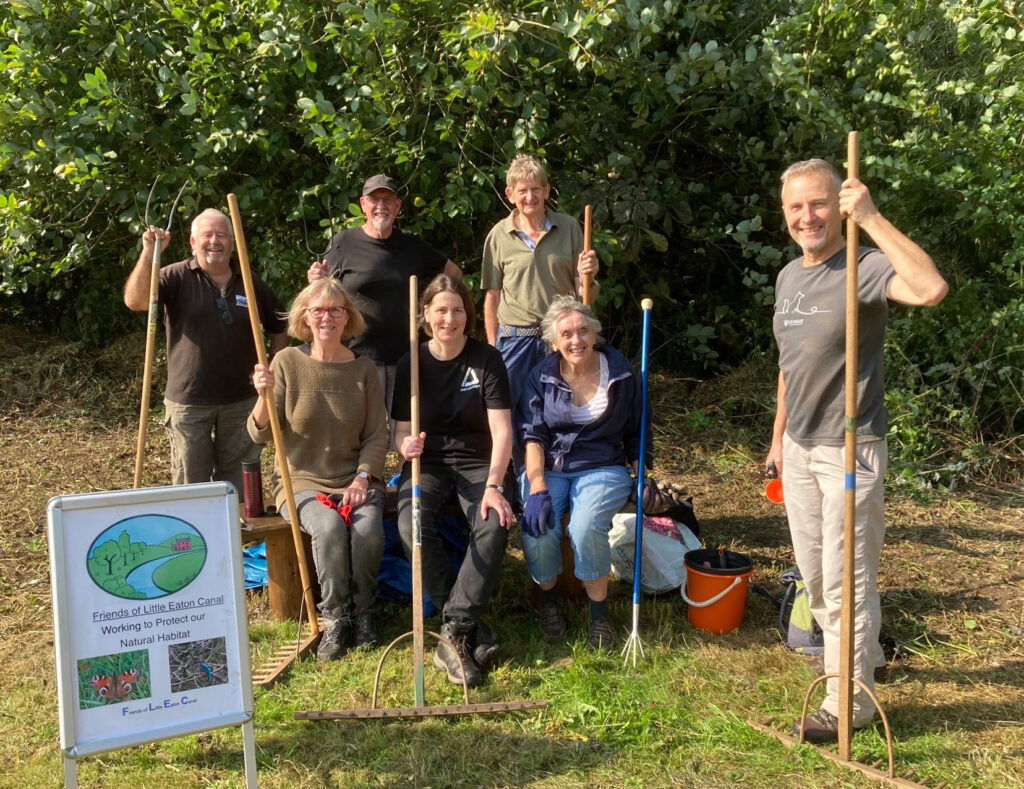 Group of people holding rakes in nature, with sign "Friends of Little Eaton Canal"