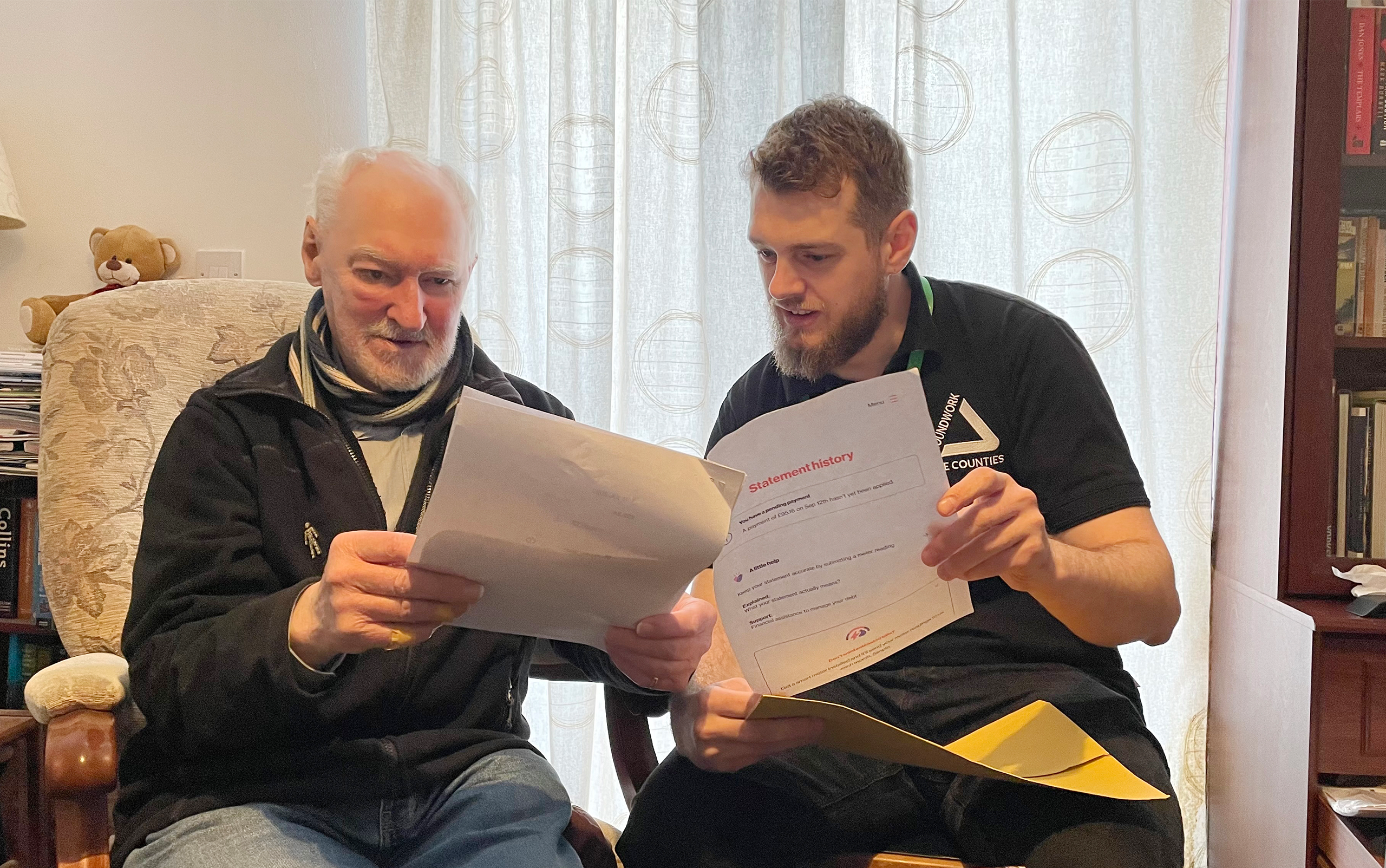 Two men looking at papers labelled 'statement history' sitting in chairs in living room.