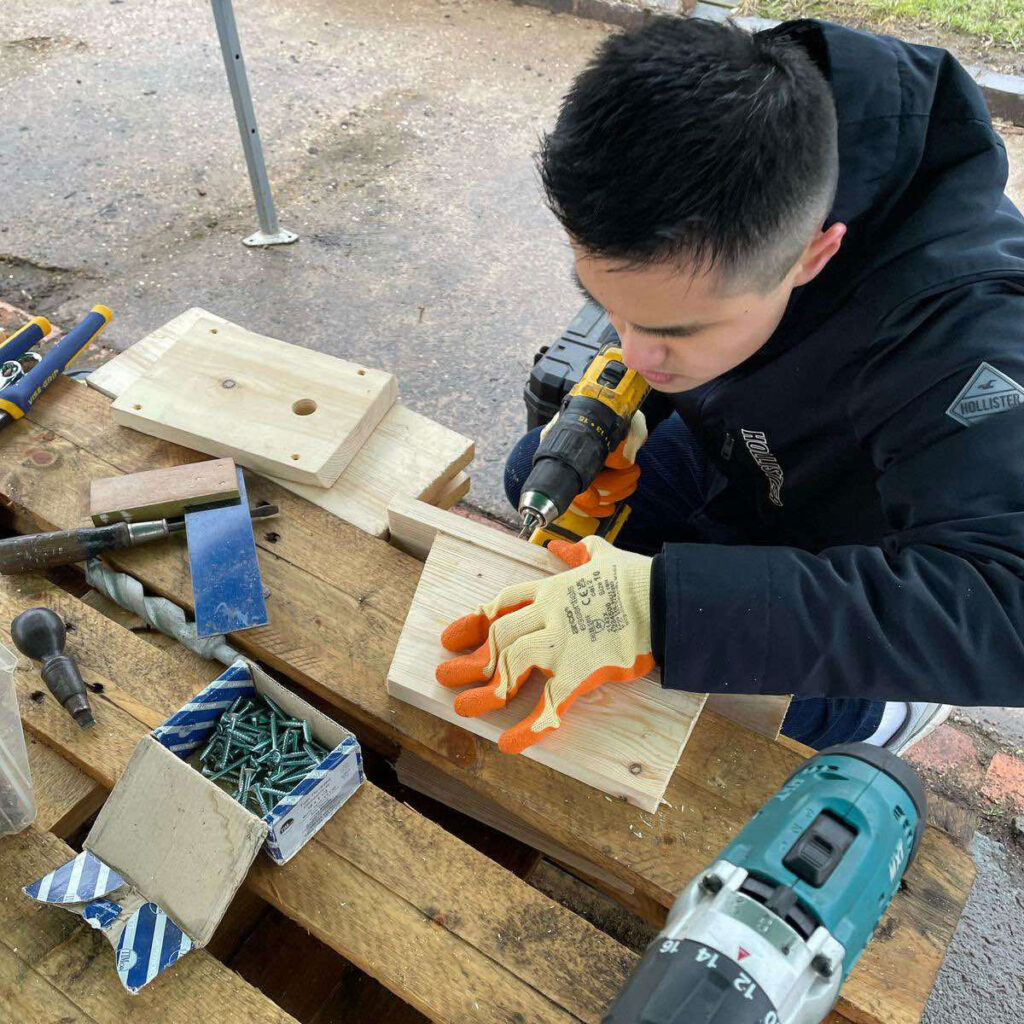 Young man with dark hair at table drilling into wood, with DIY equipment around him