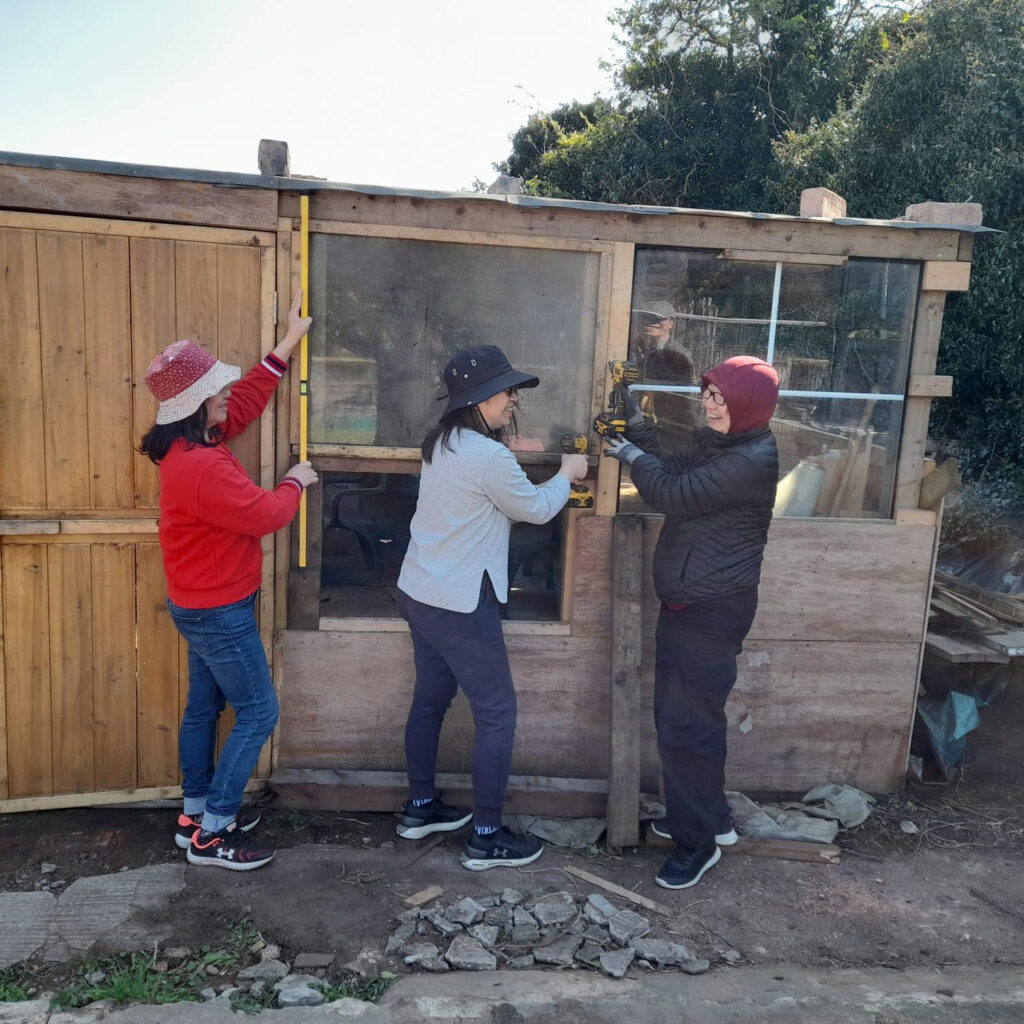 Three people holding DIY tools by shed