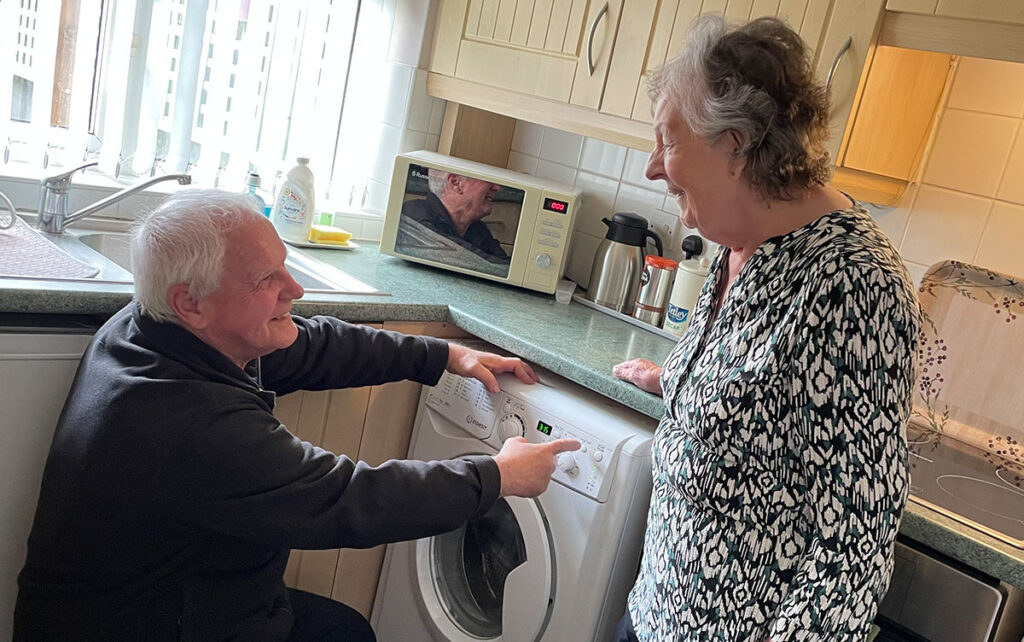 Man and woman in kitchen, man is pointing at a washing machine.