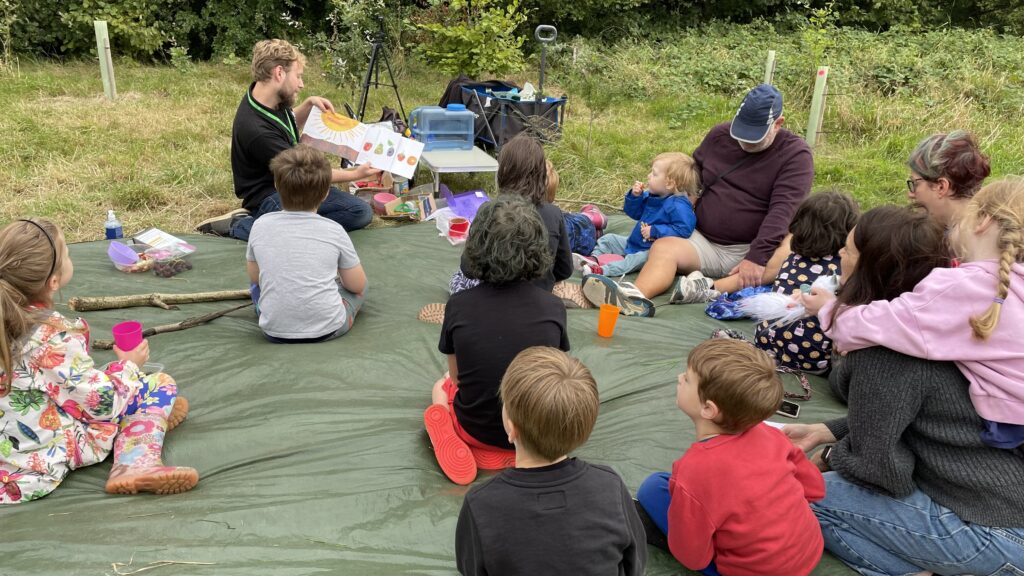 Group of children and adults sitting on tarp watching blonde man reading from a children's book.