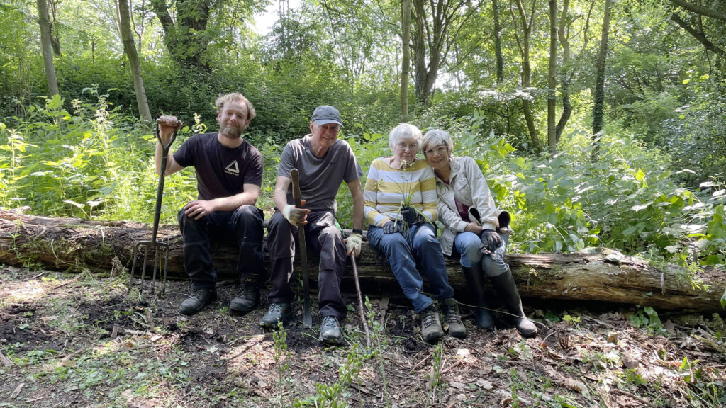 Group of two men and two women holding gardening tools, sitting on a fallen tree surrounded by trees and vegetation.