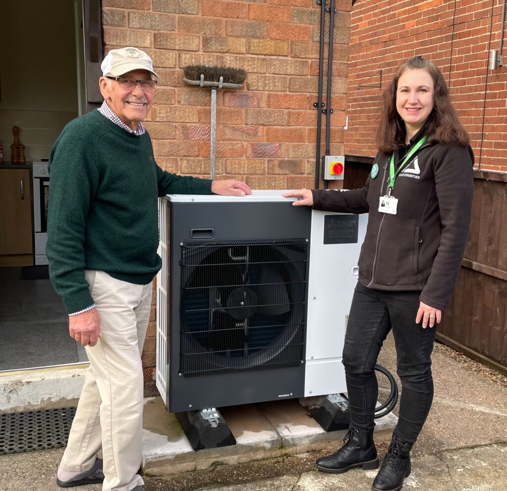 Elderly man wearing green jumper and white trousers with young woman wearing black, standing outdoors with a heat pump and smiling at the camera.