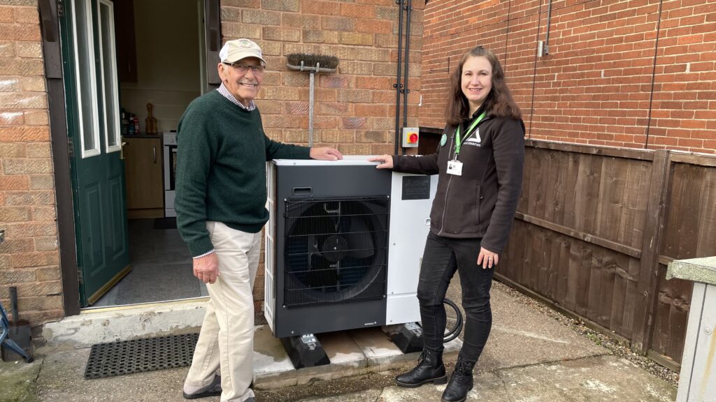 Elderly man wearing green jumper and white trousers with young woman wearing black, standing outdoors with a heat pump and smiling at the camera.