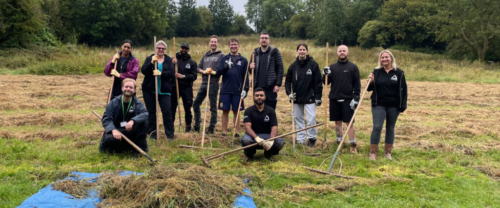 Group of people holding rakes in grassy field with blue tarpaulin full of raked grass,