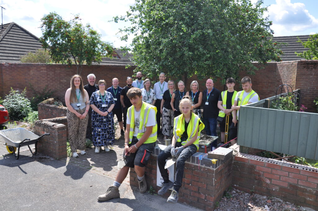 Group of men and women standing in residential area with four teenagers in yellow high visibility jackets. A wheelbarrow and construction tools are visible.