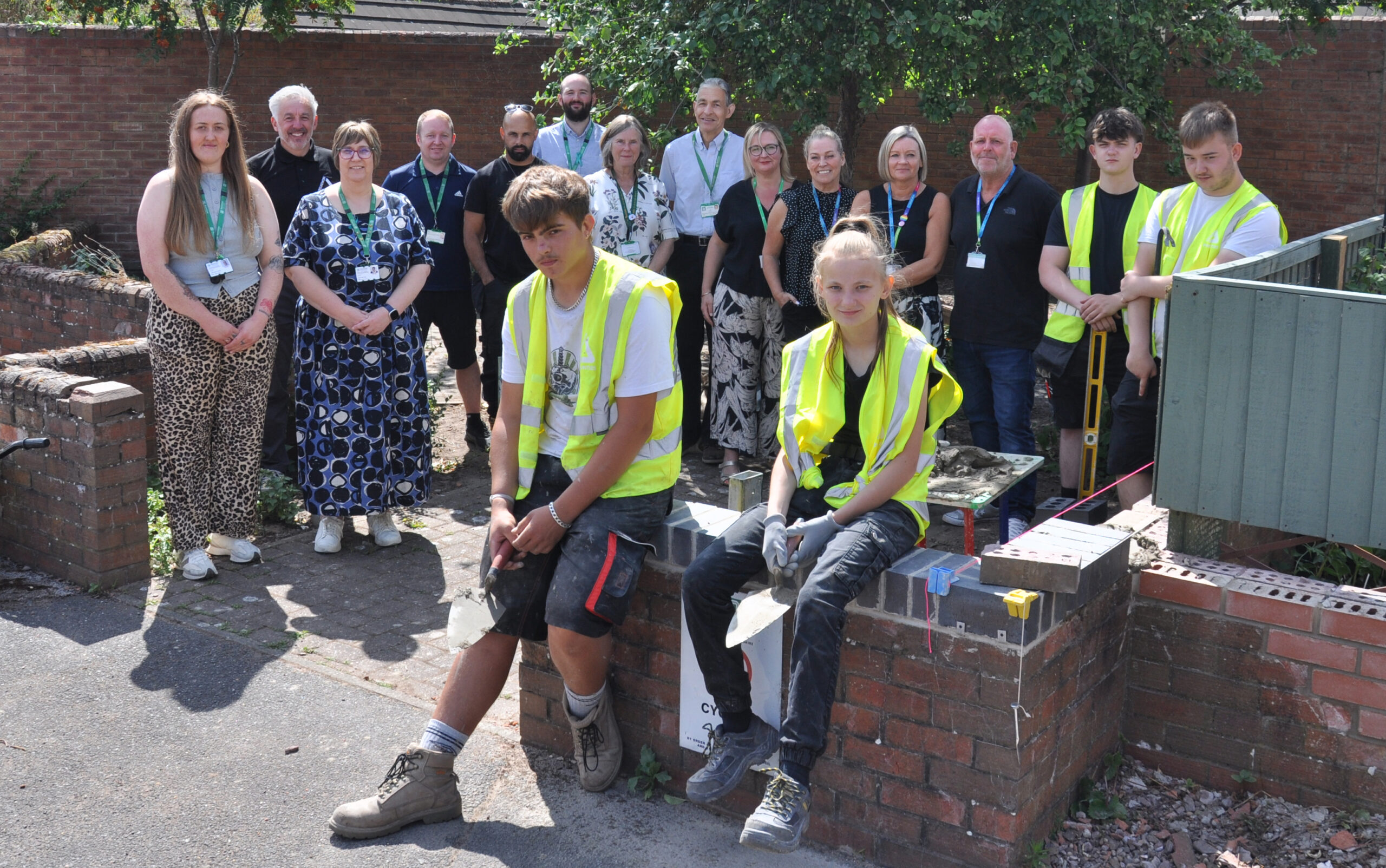 Group of men and women standing in residential area with four teenagers in yellow high visibility jackets. A wheelbarrow and construction tools are visible.