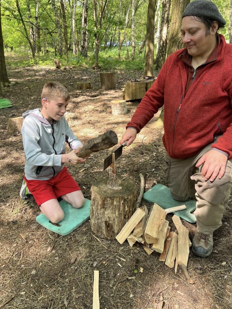 Young boy bashing large stick onto cleaver held by man in red shirt, chopping wood together in woodland area.
