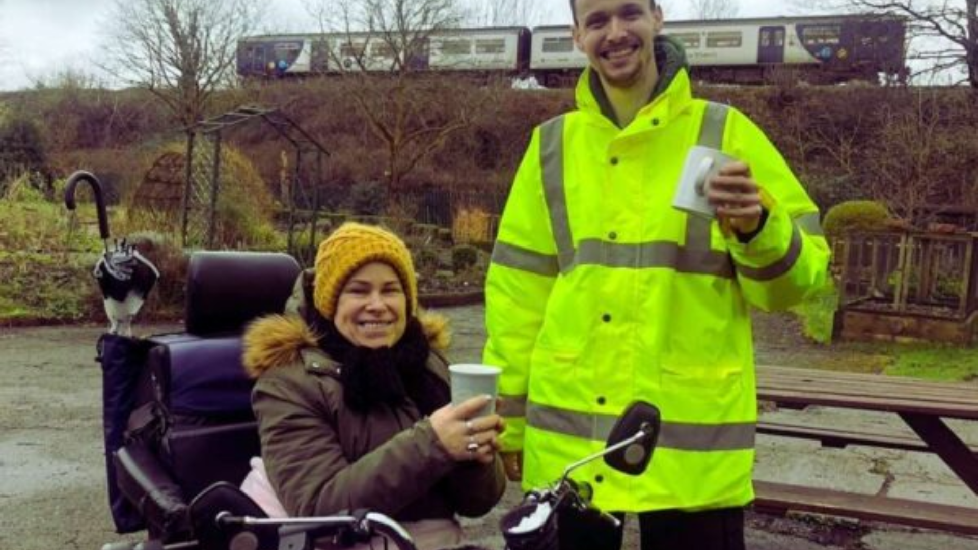 A man and woman smile holding cups of tea. They are in a community garden. The woman is in a wheelchair and wearing a wooly hat. The man is wearing a high viz jacket.