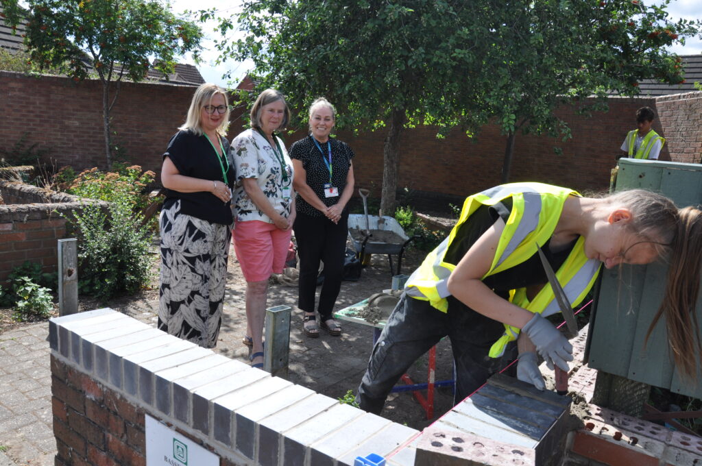Teenage girl with yellow high vis vest and trowel laying bricks on wall while three adult women watch.