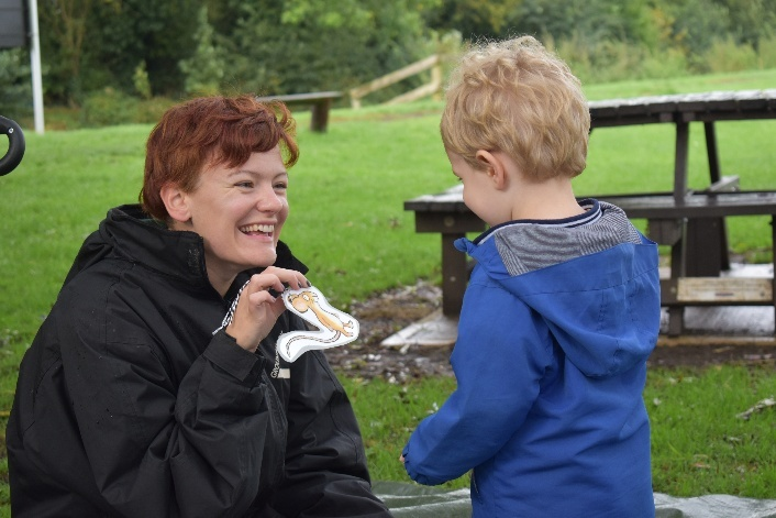 Photo of a woman kneeling down handing out a holding a cut out picture of a squierlle to a child.