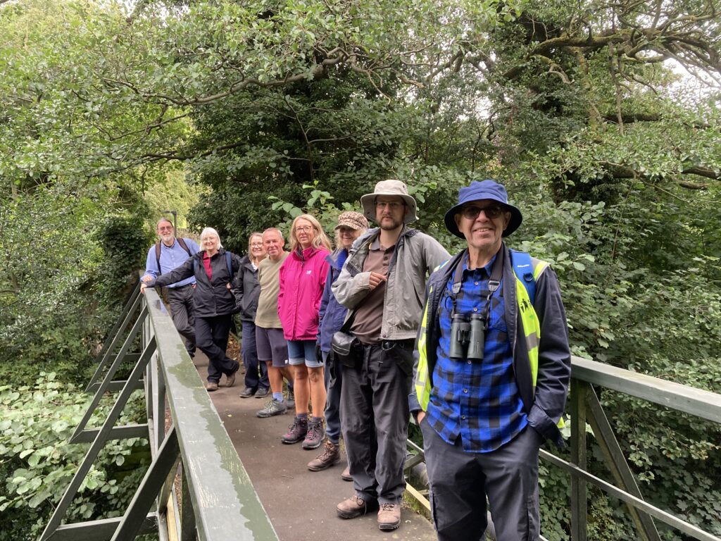 Group of people smiling at the camera standing on a bridge outdoors with trees in background.