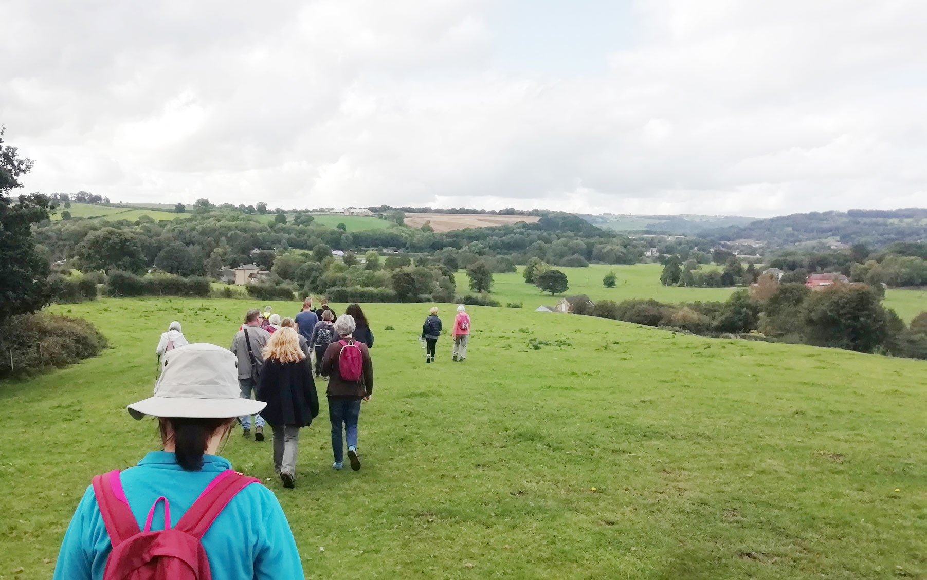 Group of people walking over grass with hills in background. Person wearing white hat, blue jacket and red backpack in foreground.