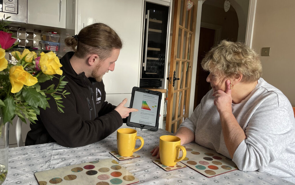Man wearing black fleece pointing at EPC information on tablet, with woman in grey top looking. They are sitting at a kitchen table with flowers and yellow cups.