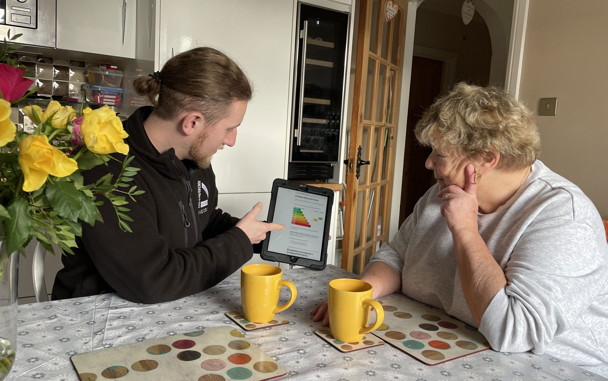 Man wearing black fleece pointing at EPC information on tablet, with woman in grey top looking. They are sitting at a kitchen table with flowers and yellow cups.