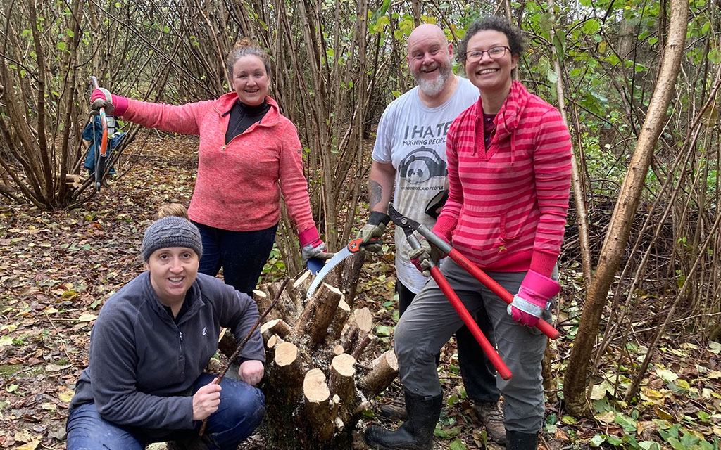 Four people holding gardening tools including saws and loopers, standing by tree stump smiling at the camera in wooded outdoor area.