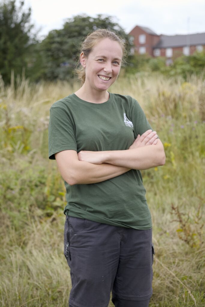 Photo of a woman in a green Groundwork shirt stands smiling with her arms crossed outside an allotment centre.