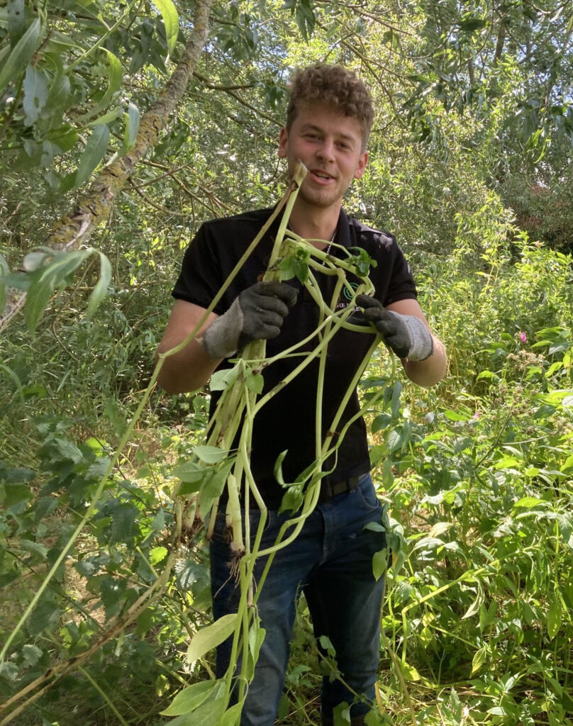 Young man wearing black top and garden gloves holding plants in green outdoor space.