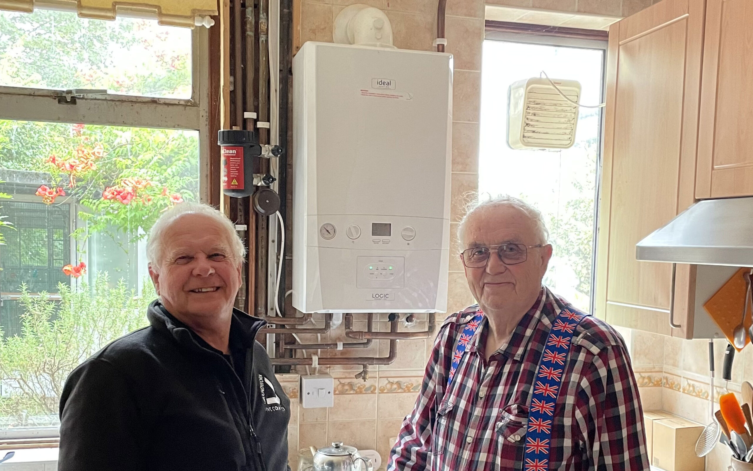Two white-haired men standing in kitchen smiling at camera with brand new combi-boiler in background.