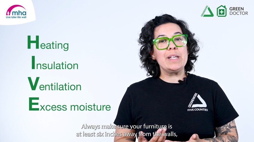 Screenshot of video: Woman with black hair and green glasses wearing black T-shirt with white logo in front of white background. Green text "Heating, Insulation, Ventilation, Excess Moisture" and white subtitles at the bottom. In the top left corner is a pink and blue logo, top right corner has green and black Groundwork and Green Doctor logos.