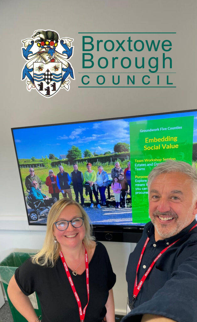 Blonde woman and man with white hair and neat beard wearing red lanyards in front of TV screen showing group of people, green box and white and yellow text "Groundwork Five Counties - Embedding Social Value". The Broxtowe Borough Council logo iis superimposed at the top of the photo.