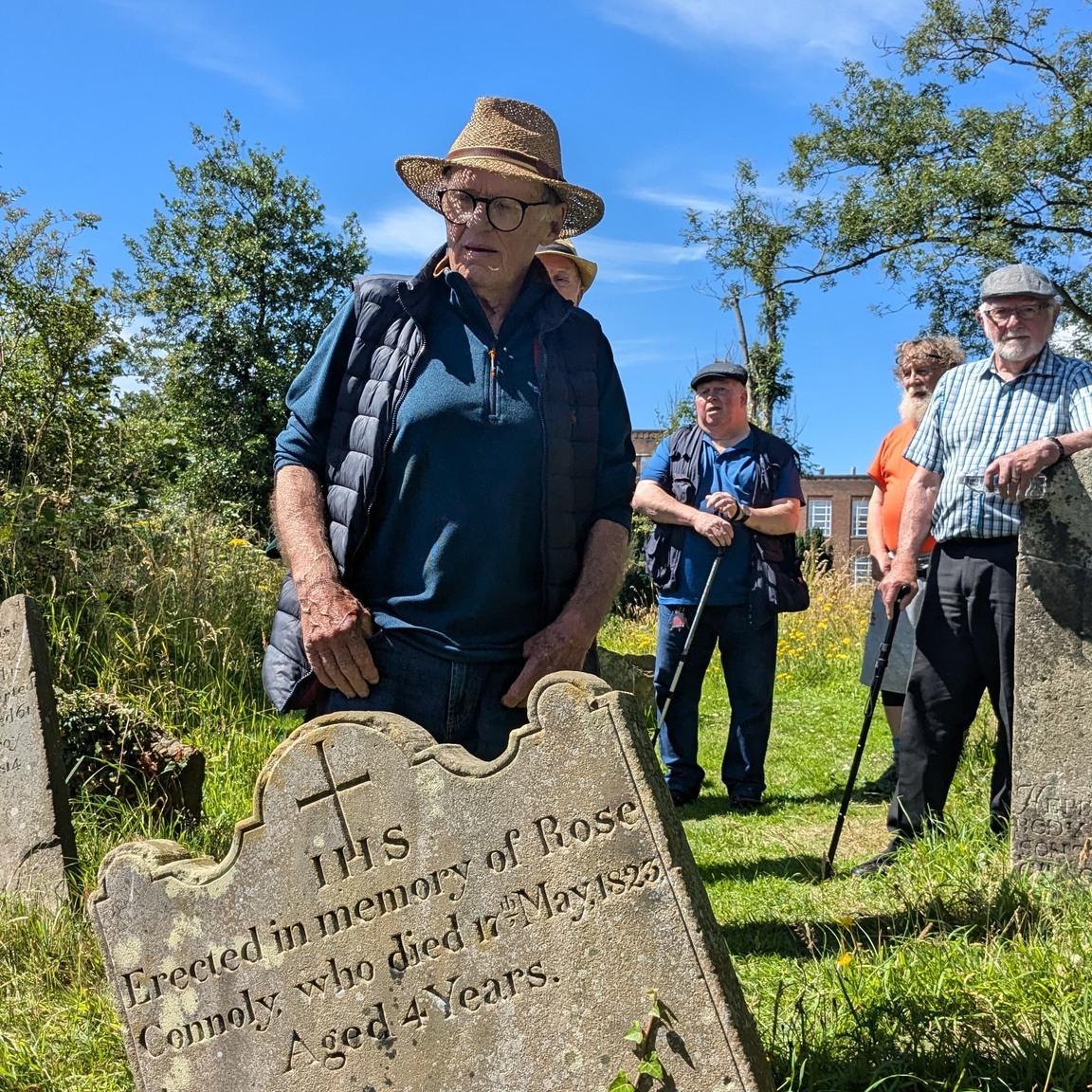 A group of men on a sunny day beside a very old gravestone