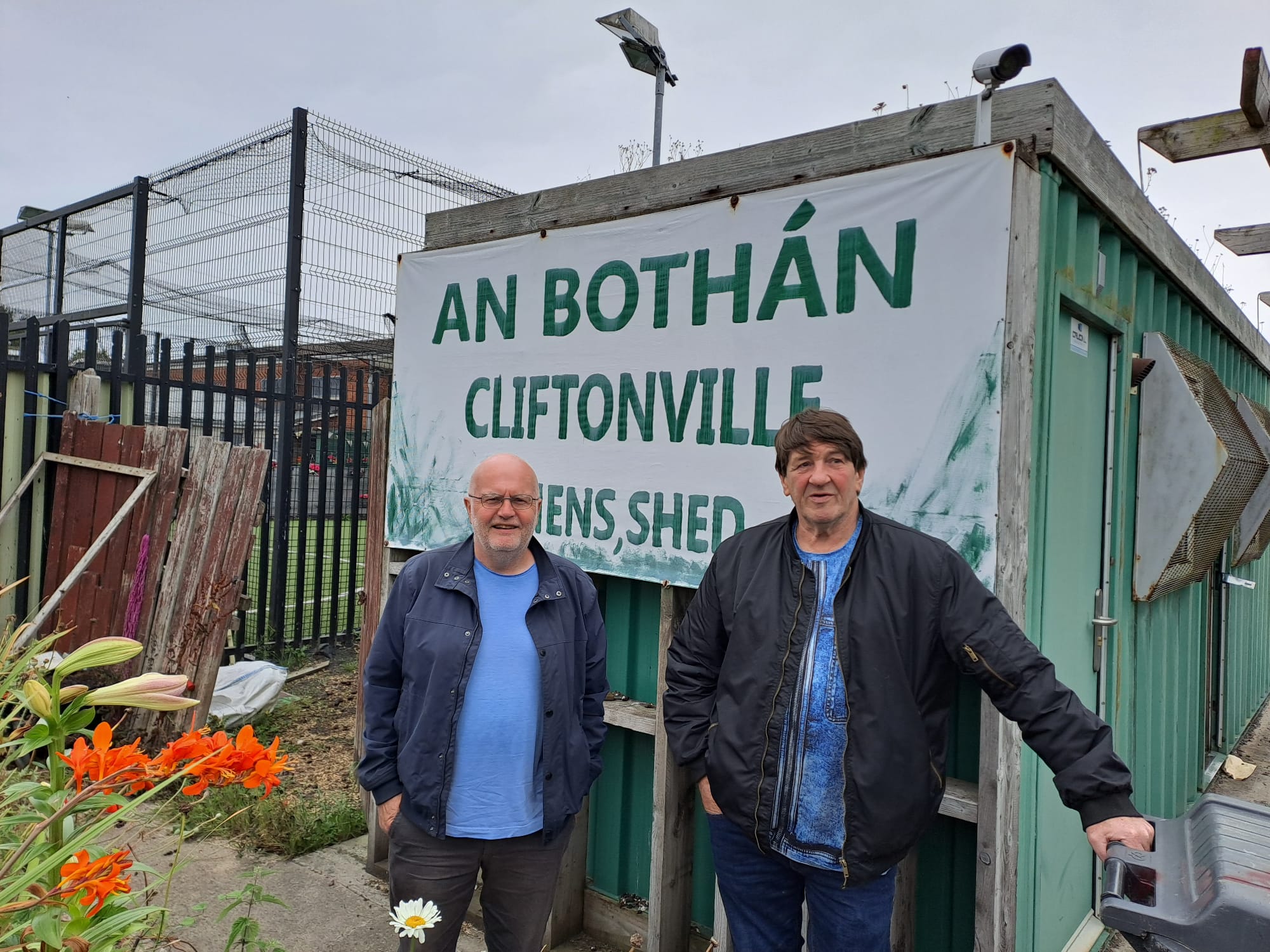 Two men beside some flowers and a shipping container with a sign saying "An Bothan Cliftonville Men's Shed"