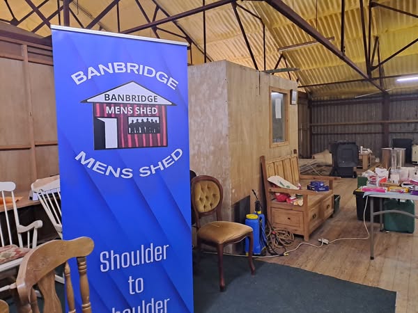 A plyboard room build within a barn, a scattering of chairs and a pop banner reading "Banbridge Men's Shed"