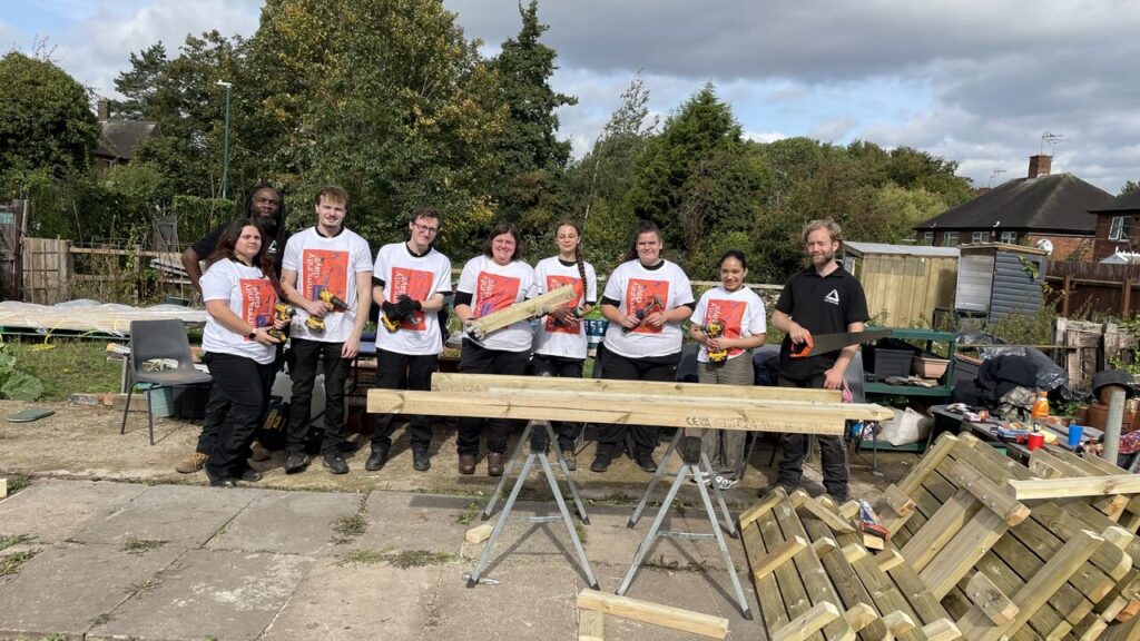 Group of people wearing white and black T-shirts in outdoor space with planks of wood in foreground. People are smiling at the camera and holding power tools