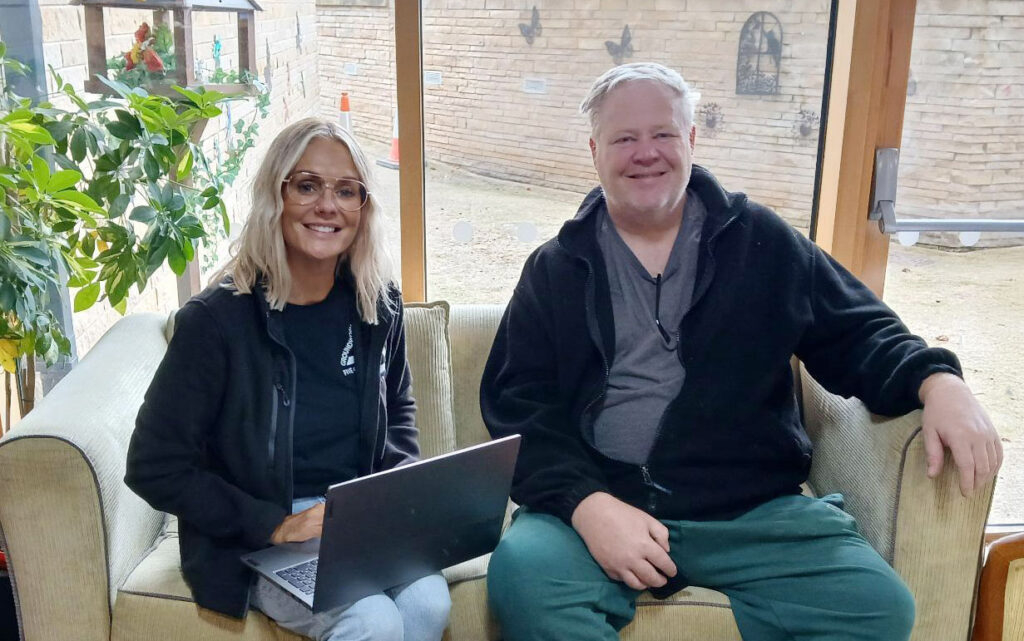 Blonde woman and older man sitting on beige sofa smiling at the camera.