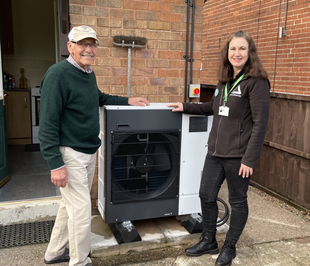 Elderly man wearing green jumper and white trousers with young woman wearing black, standing outdoors with a heat pump and smiling at the camera.