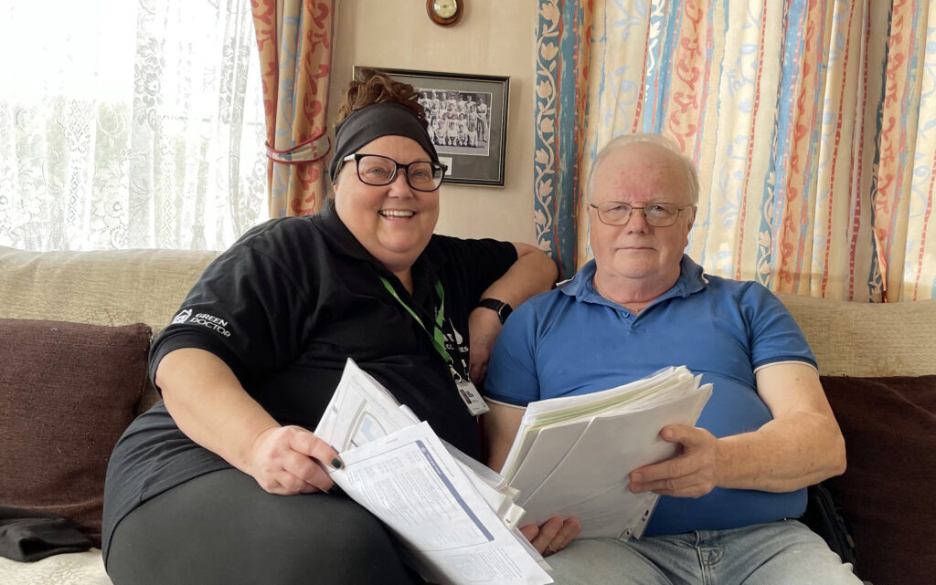 Woman wearing black top and bandana sitting next to elderly man in blue shirt. Both are holding paperwork and are smiling at the camera.