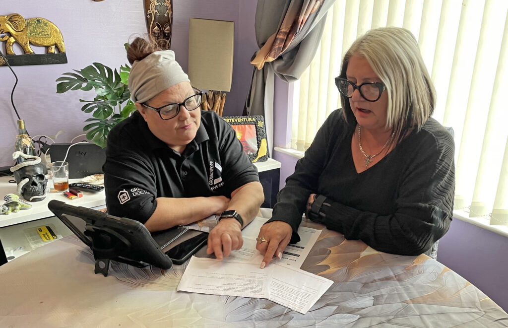 Two women looking at bills laid out on table in room with purple walls and knickknacks in the background.