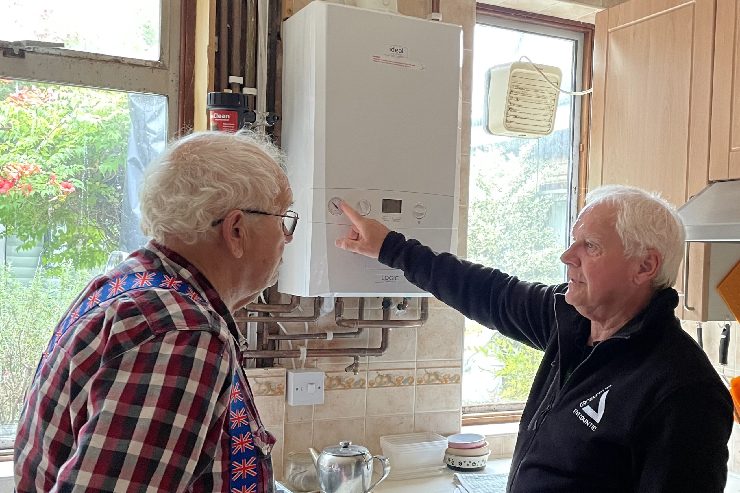 Man wearing black fleece pointing at gauge on combi boiler, looking at ederly man in plaid shirt in cosy kitchen.