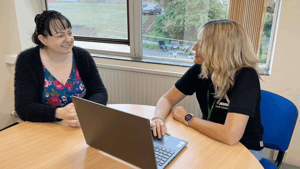 Dark haired woman and blonde woman sitting at desk, talking with laptop.