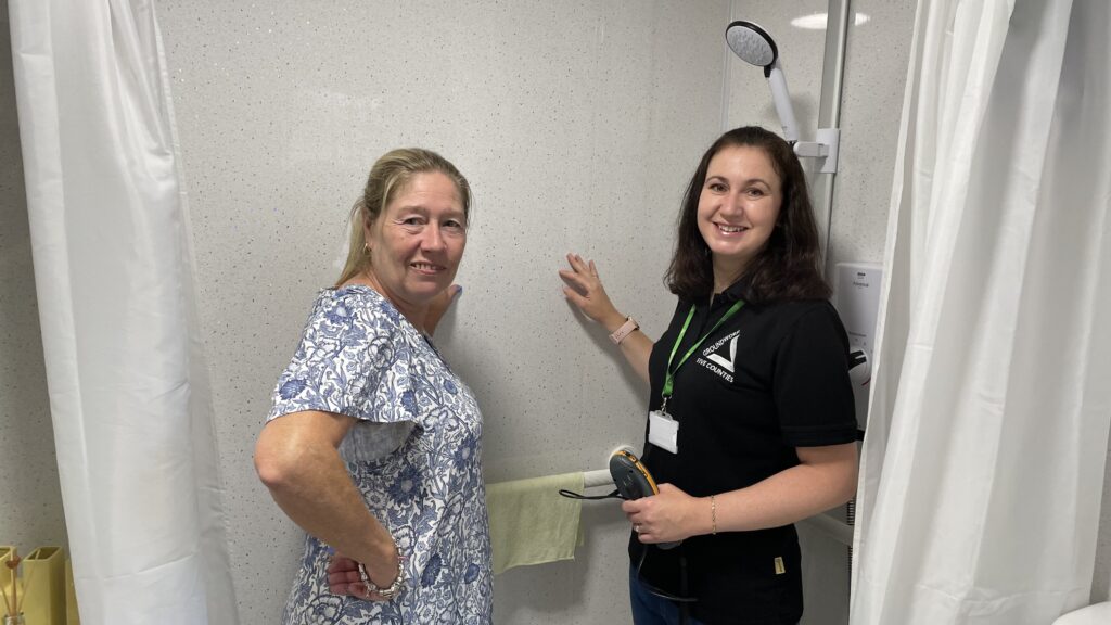 Two women standing in bathroom touching wall and smiling at the camera.