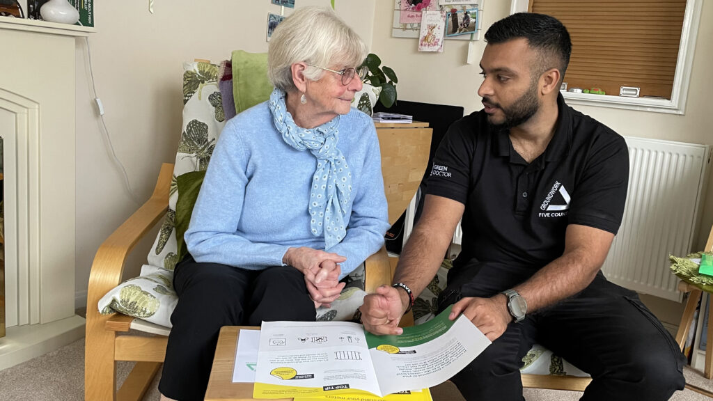 Elderly woman wearing black top speaking with man wearing black, sitting in chairs in cosy room looking at booklet on table.