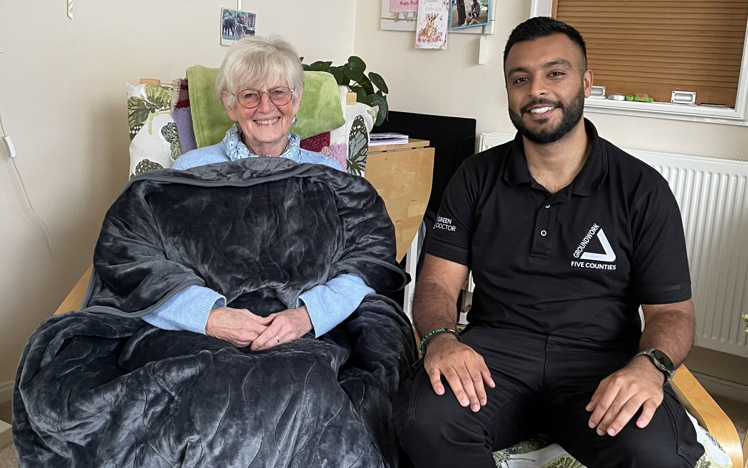 Elderly woman under dark grey blanket sitting in a cosy room next to man wearing black clothing with white Groundwork Five Counties logo.