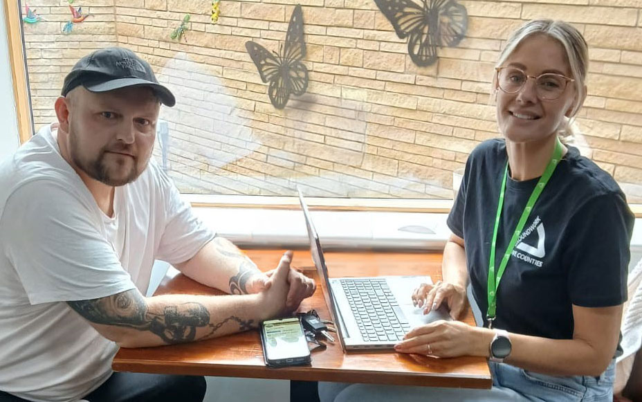 Man wearing black cap and white T-shirt sitting at wooden table with blonde woman wearing black T-shirt and green lanyard.